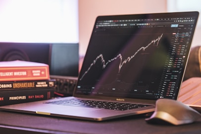 A laptop displaying a stock market graph with a rising trend is placed on a desk. Next to the laptop are several books stacked on each other, with titles related to investing. A mouse is positioned to the right of the laptop.