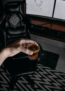 A hand holding a small glass filled with a brown liquid, possibly a beverage or espresso shot, in front of a black leather gaming chair. The background includes a computer setup with monitors and a keyboard, sitting on a wooden desk. The floor has a black and white zigzag patterned rug.