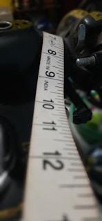 A close-up view of a measuring tape extended across a surface, with numbers and markings clearly visible. The tape is primarily white with black numbering and the text 'Made in India' is printed on it. Parts of tools and possibly machinery are visible in the blurred background.