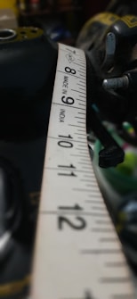A close-up view of a measuring tape extended across a surface, with numbers and markings clearly visible. The tape is primarily white with black numbering and the text 'Made in India' is printed on it. Parts of tools and possibly machinery are visible in the blurred background.
