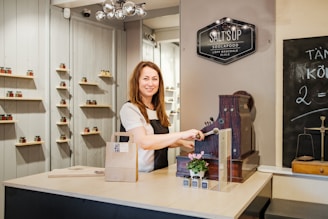 A woman stands behind a counter with a vintage cash register, smiling while operating it. The counter holds a small potted plant and some small bottles. Shelves on the background wall display jars with various colors. A chalkboard on the right side shows some writing. A signboard above reads 'SALT'S UP SOOLAPOOD'.