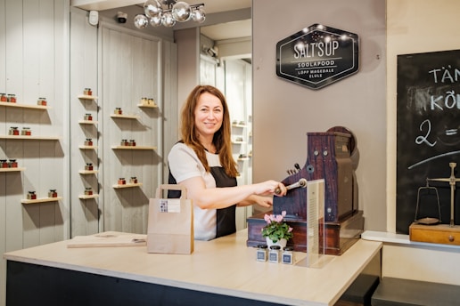 A woman stands behind a counter with a vintage cash register, smiling while operating it. The counter holds a small potted plant and some small bottles. Shelves on the background wall display jars with various colors. A chalkboard on the right side shows some writing. A signboard above reads 'SALT'S UP SOOLAPOOD'.