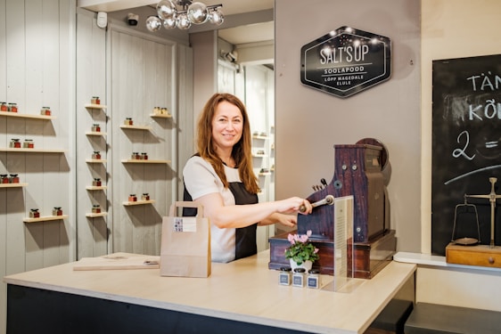 A woman stands behind a counter with a vintage cash register, smiling while operating it. The counter holds a small potted plant and some small bottles. Shelves on the background wall display jars with various colors. A chalkboard on the right side shows some writing. A signboard above reads 'SALT'S UP SOOLAPOOD'.