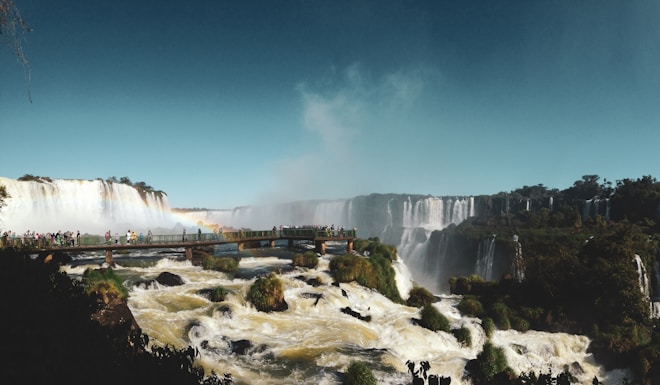 A vast and powerful waterfall cascades over a rocky cliff, surrounded by lush greenery. A bridge filled with people offers a vantage point to view the falls, with mist rising prominently in the background.