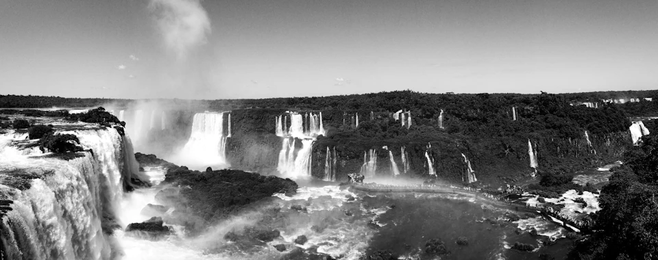 The image depicts a breathtaking view of the Iguazu Falls, one of the world's largest waterfalls. The falls are captured from an elevated perspective, showcasing their grandeur and power. The waterfall is situated on the left side of the frame, with its white waters cascading down into a pool below. The surrounding landscape features lush green trees and vegetation, creating a vibrant contrast against the clear blue sky above.
