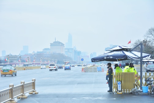 A wide, urban street scene with several vehicles including cars and taxis driving down the road. On the right side, there is a group of police officers wearing yellow reflective jackets standing near a small kiosk with a police sign. The background features modern buildings under a gray, overcast sky. Flags are lined along the street, and a red national flag is visible flying in the distance.