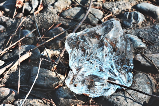 A clear block of ice lies on a rough surface of uneven, gray stones. Dry twigs are scattered across the stones, adding a natural texture. The ice captures sunlight, creating reflections and highlighting its transparent quality.