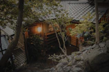 A traditional Japanese house is nestled among lush green trees. The building features wooden walls, a tiled roof, and a softly glowing lantern at the entrance. Stones and foliage surround the structure, enhancing its serene and natural setting.