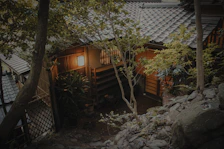 A traditional Japanese house is nestled among lush green trees. The building features wooden walls, a tiled roof, and a softly glowing lantern at the entrance. Stones and foliage surround the structure, enhancing its serene and natural setting.