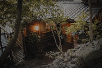 A traditional Japanese house is nestled among lush green trees. The building features wooden walls, a tiled roof, and a softly glowing lantern at the entrance. Stones and foliage surround the structure, enhancing its serene and natural setting.