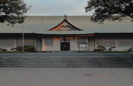 A traditional Japanese-style building with a symmetrical facade and prominent roof, surrounded by neatly trimmed greenery. The structure features white walls with vertical window slats and a centered entrance, accentuated by warm lighting from within. The sky is slightly overcast, and the scene includes trees framing the view.
