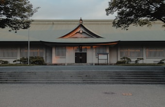 A traditional Japanese-style building with a symmetrical facade and prominent roof, surrounded by neatly trimmed greenery. The structure features white walls with vertical window slats and a centered entrance, accentuated by warm lighting from within. The sky is slightly overcast, and the scene includes trees framing the view.