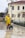 A person wearing a yellow raincoat, protective pants, and a mask is engaged in street cleaning. They are pushing a green trash bin while holding a broom. The scene takes place on a wet pavement in front of a light-colored building with arched windows and a tiled roof. Leafless trees and a street lamp are also visible in the background.