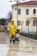 A person wearing a yellow raincoat, protective pants, and a mask is engaged in street cleaning. They are pushing a green trash bin while holding a broom. The scene takes place on a wet pavement in front of a light-colored building with arched windows and a tiled roof. Leafless trees and a street lamp are also visible in the background.