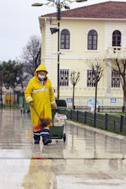 A person wearing a yellow raincoat, protective pants, and a mask is engaged in street cleaning. They are pushing a green trash bin while holding a broom. The scene takes place on a wet pavement in front of a light-colored building with arched windows and a tiled roof. Leafless trees and a street lamp are also visible in the background.
