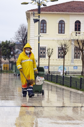 A person wearing a yellow raincoat, protective pants, and a mask is engaged in street cleaning. They are pushing a green trash bin while holding a broom. The scene takes place on a wet pavement in front of a light-colored building with arched windows and a tiled roof. Leafless trees and a street lamp are also visible in the background.