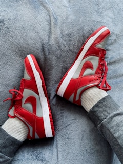 A pair of red and white sneakers are worn by a person lying on a soft, textured gray surface. The person appears to be dressed in gray sweatpants and cream-colored socks.