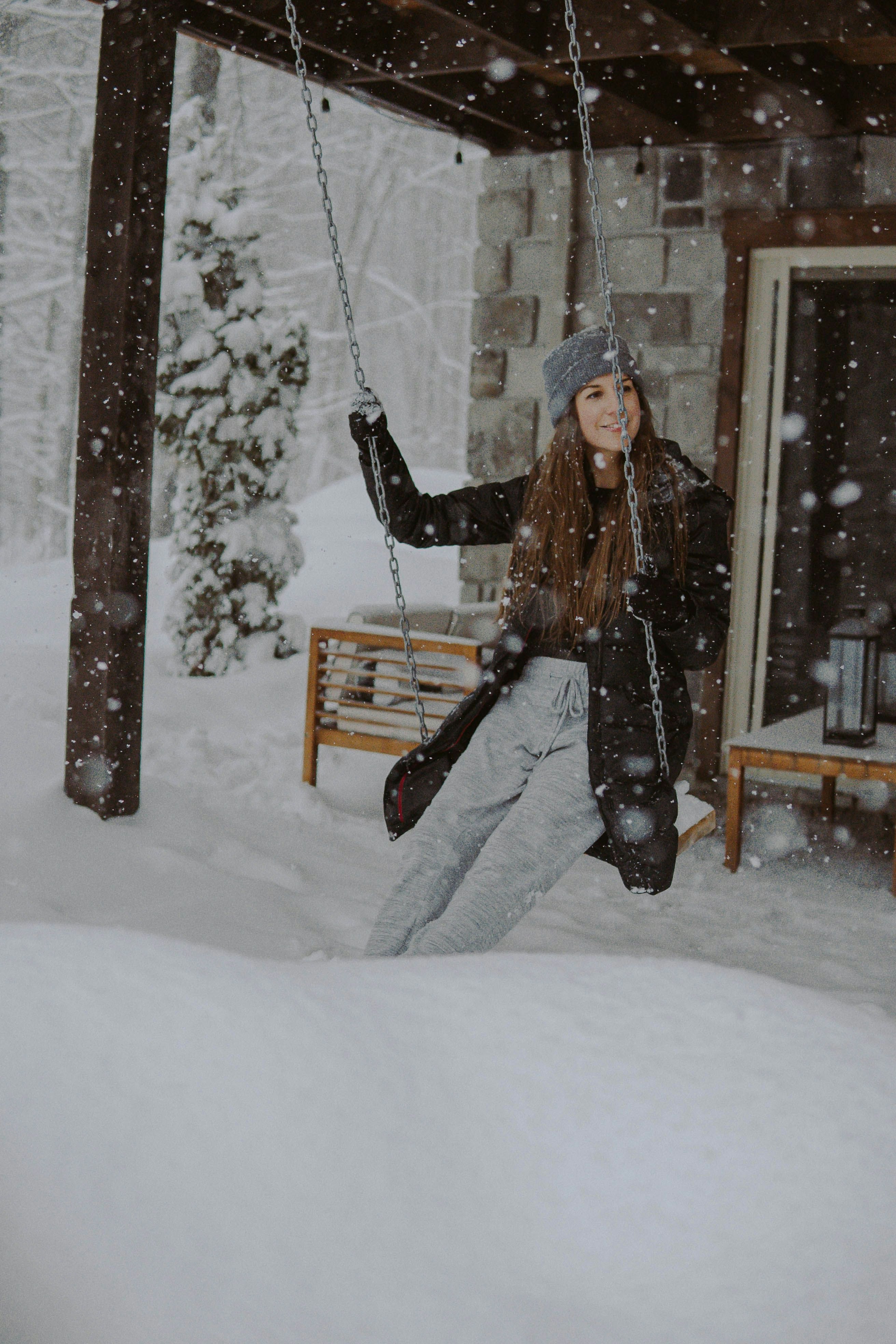 woman in black jacket and black pants sitting on brown wooden swing covered with snow