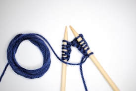 Two wooden knitting needles are intertwined with dark blue yarn against a plain white background. One end of the yarn is coiled into a neat circle while the other extends, wrapping around the needles in a simple knitting pattern.
