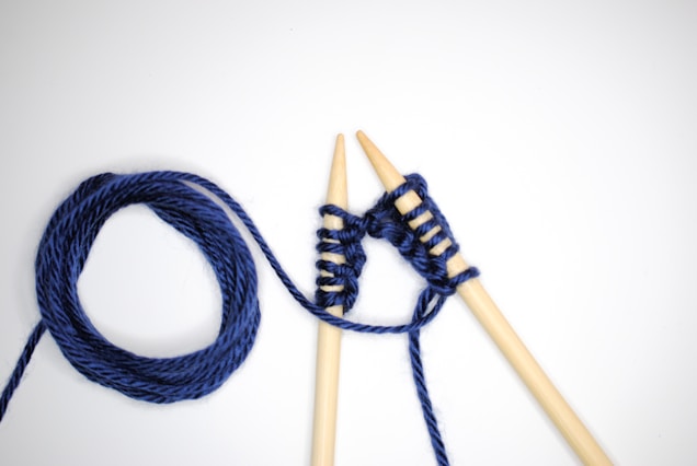 Two wooden knitting needles are intertwined with dark blue yarn against a plain white background. One end of the yarn is coiled into a neat circle while the other extends, wrapping around the needles in a simple knitting pattern.
