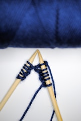 Wooden knitting needles are being used to knit with dark blue yarn. The background is a soft gradient of white to an intense blue tone, likely comprised of unfocused blue yarn.