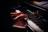 Hands playing on a grand piano with visible brand markings, with sheet music placed above the keys. The lighting is soft, highlighting the player's hands and the piano keys.