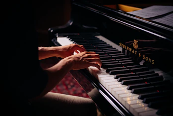 Hands playing on a grand piano with visible brand markings, with sheet music placed above the keys. The lighting is soft, highlighting the player's hands and the piano keys.