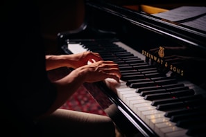 Hands playing on a grand piano with visible brand markings, with sheet music placed above the keys. The lighting is soft, highlighting the player's hands and the piano keys.