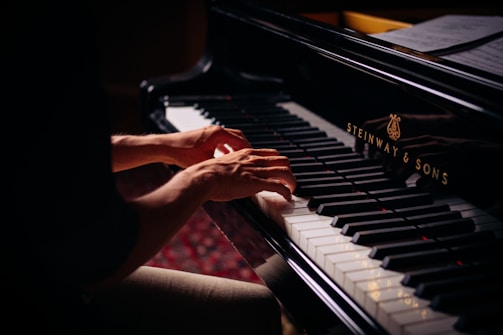 Hands playing on a grand piano with visible brand markings, with sheet music placed above the keys. The lighting is soft, highlighting the player's hands and the piano keys.