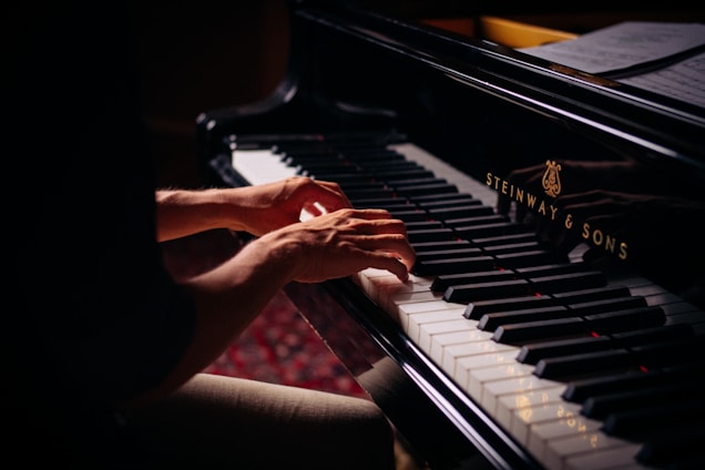 Hands playing on a grand piano with visible brand markings, with sheet music placed above the keys. The lighting is soft, highlighting the player's hands and the piano keys.