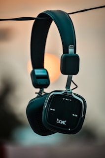 A stylish pair of black over-ear headphones is hanging outdoors against a blurred background with a warm sunset glow. The headphones have a sleek design with soft cushioning and visible control buttons on the earpiece.