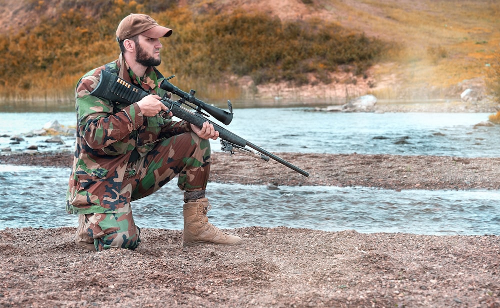 Mann in grüner und brauner Tarnjacke, der tagsüber ein Gewehr in der Nähe des Gewässers hält