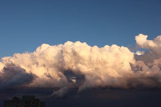 Large, fluffy clouds in varying shades of white and gray dominate the sky against a backdrop of clear blue. A small section of building rooftop is visible at the bottom, slightly shadowed beneath the clouds.
