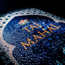 A close-up view of a blue surface covered with water droplets. The words 'Taj Mahal' are prominently displayed in bold, golden letters against the blue background. The surface appears textured, creating a visually striking pattern due to the light interacting with the droplets.