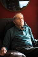 An elderly man sits in a cushioned armchair, wearing a teal long-sleeve polo shirt. The background features a deep red wall with a mirror reflecting part of the room.