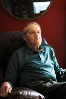 An elderly man sits in a cushioned armchair, wearing a teal long-sleeve polo shirt. The background features a deep red wall with a mirror reflecting part of the room.