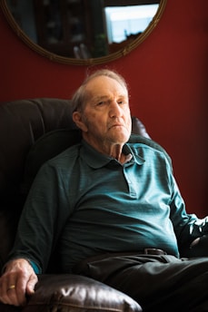 An elderly man sits in a cushioned armchair, wearing a teal long-sleeve polo shirt. The background features a deep red wall with a mirror reflecting part of the room.