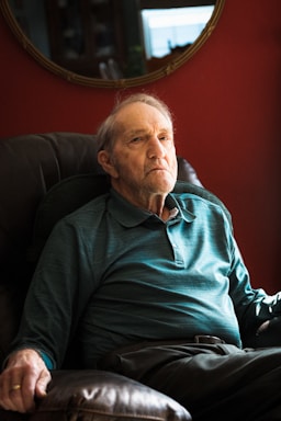 An elderly man sits in a cushioned armchair, wearing a teal long-sleeve polo shirt. The background features a deep red wall with a mirror reflecting part of the room.