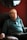An elderly man sits in a cushioned armchair, wearing a teal long-sleeve polo shirt. The background features a deep red wall with a mirror reflecting part of the room.