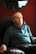 An elderly man sits in a cushioned armchair, wearing a teal long-sleeve polo shirt. The background features a deep red wall with a mirror reflecting part of the room.