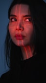 A woman with long dark hair gazes off to the side, illuminated by red laser lines that cast a grid pattern across her face. Her expression is contemplative, set against a dark background, enhancing the dramatic lighting.