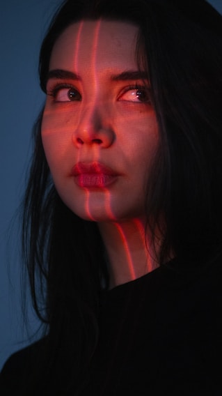 A woman with long dark hair gazes off to the side, illuminated by red laser lines that cast a grid pattern across her face. Her expression is contemplative, set against a dark background, enhancing the dramatic lighting.