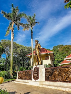 A bronze statue of a person in uniform stands prominently in front of a building with a red-tiled roof surrounded by lush greenery. Tall palm trees tower next to the statue, and there are intricate engravings on bronze panels beside it. The scene is set against a bright blue sky with scattered clouds.