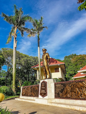 A bronze statue of a person in uniform stands prominently in front of a building with a red-tiled roof surrounded by lush greenery. Tall palm trees tower next to the statue, and there are intricate engravings on bronze panels beside it. The scene is set against a bright blue sky with scattered clouds.