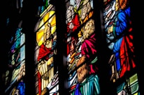 Intricate stained glass artwork depicting religious figures in vivid colors. The scene includes several people dressed in robes, surrounded by symbolic elements and radiant hues. Detailed craftsmanship is evident in the expressions and gestures of the figures portrayed.
