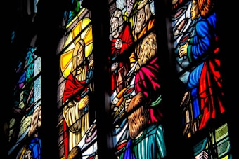 Intricate stained glass artwork depicting religious figures in vivid colors. The scene includes several people dressed in robes, surrounded by symbolic elements and radiant hues. Detailed craftsmanship is evident in the expressions and gestures of the figures portrayed.