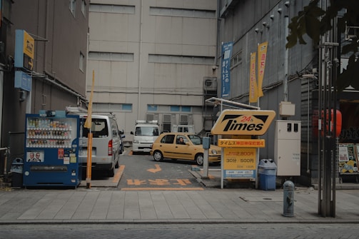 A small urban parking lot with two vehicles parked, one white van and one yellow car. There's a vending machine on the left, a prominently displayed 'Times' parking sign with pricing details in the foreground, and several banners with Japanese text attached to the buildings. The area is enclosed by tall, grey buildings with windows and some visible street infrastructure.