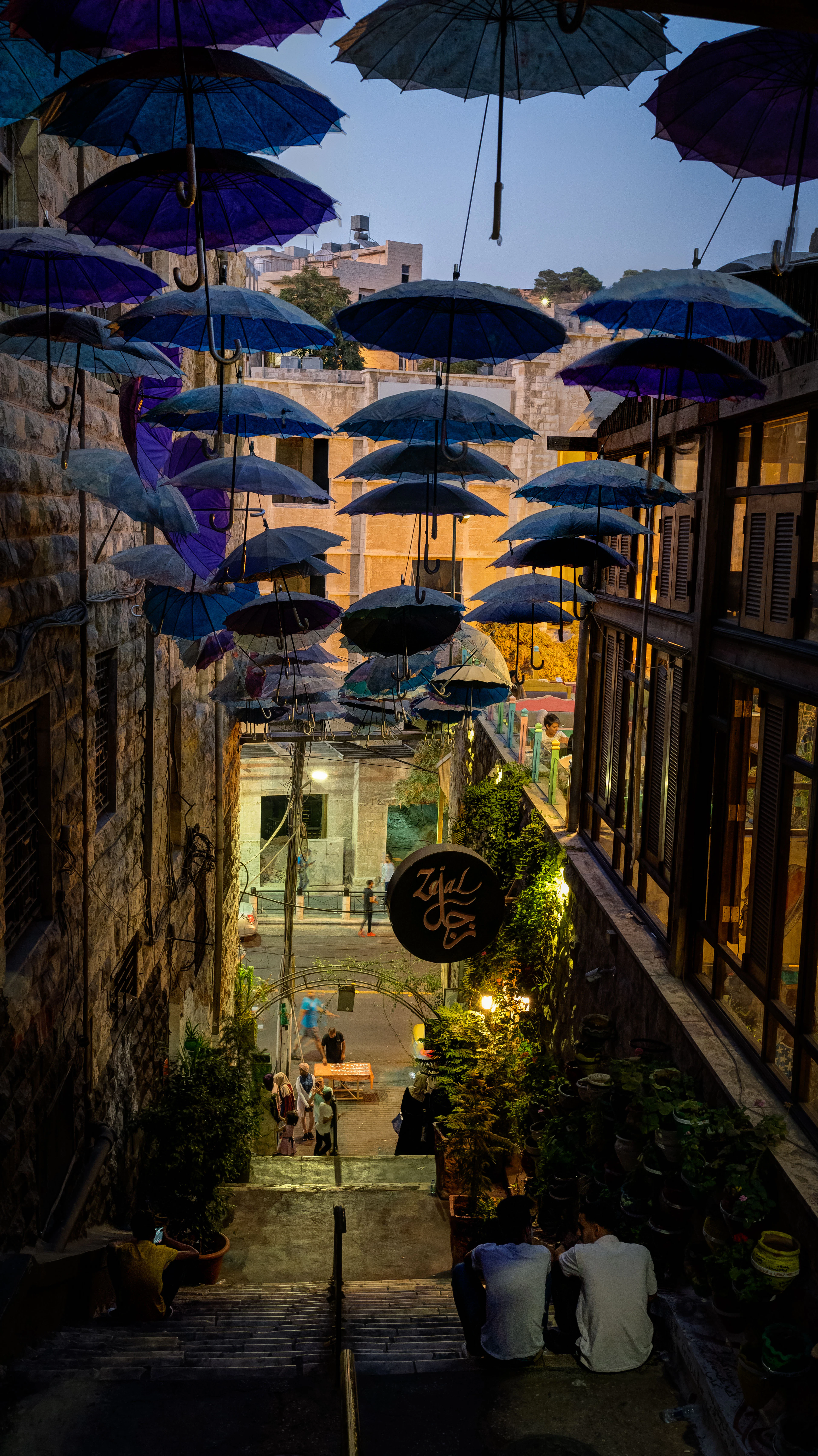 Blue and violet umbrellas strung above a stone stairway in downtown Amman at dusk, warm restaurant lights and greenery below, and people sitting out on the steps in the evening.
