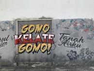 Graffiti art on a wall featuring bold text in yellow, red, and white that says 'Gomo Kelate Gomo!' with a cracked effect. Floral patterns and additional text 'Tanah Airku' are also drawn in decorative fonts on the sides. The background seems to be an old, weathered wall with some stains and cracks.