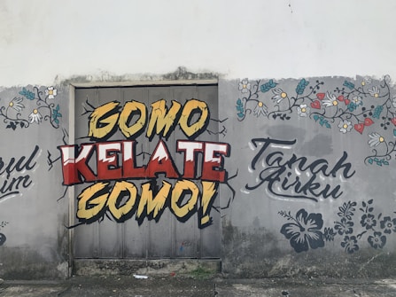 Graffiti art on a wall featuring bold text in yellow, red, and white that says 'Gomo Kelate Gomo!' with a cracked effect. Floral patterns and additional text 'Tanah Airku' are also drawn in decorative fonts on the sides. The background seems to be an old, weathered wall with some stains and cracks.